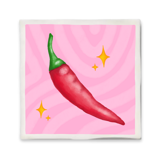 Decoratief keramisch tegeltje met een illustratie van een rode peper op een roze achtergrond met fonkelende details. Formaat 10x10 cm, voorzien van haakje om op te hangen.